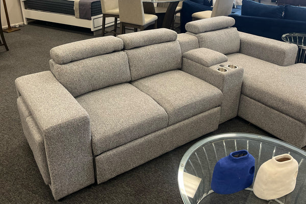 Loveseat moderno en color gris claro con tapizado texturizado, reposacabezas ajustables y consola central con dos portavasos. Exhibido en una tienda de muebles junto a una chaise longue a juego y una mesa auxiliar con cubierta de vidrio.