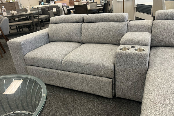 Loveseat moderno en color gris claro con tapizado texturizado, reposacabezas ajustables y consola central integrada con dos portavasos metálicos. Exhibido en una tienda de muebles junto a una chaise longue modular a juego.