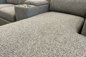 Primer plano de la chaise longue de un sofá seccional gris claro con tapizado texturizado. Se aprecia la superficie amplia del asiento y, al fondo, una consola integrada con portavasos metálicos a juego.