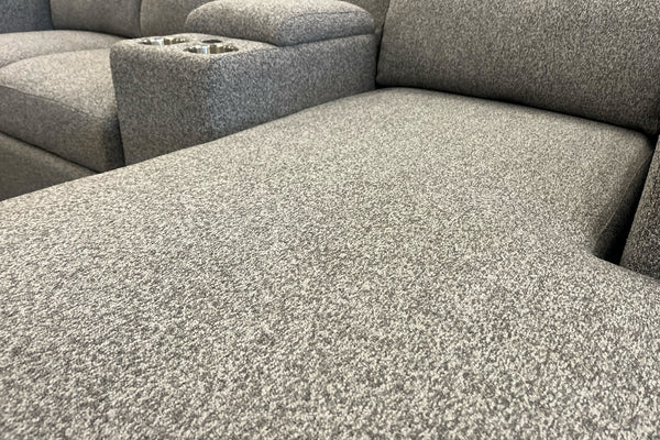 Primer plano de la chaise longue de un sofá seccional gris claro con tapizado texturizado. Se aprecia la superficie amplia del asiento y, al fondo, una consola integrada con portavasos metálicos a juego.