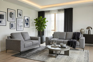 Sala moderna con sofá y loveseat tapizados en tela gris claro, mesa de centro redonda, alfombra en tonos gris y beige, cuadros abstractos en la pared y grandes ventanas con cortinas translúcidas y oscuras.
