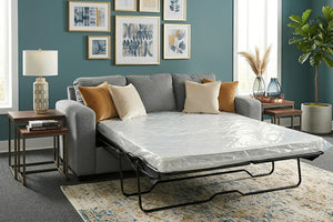 Sofá cama tapizado en tela gris con mattress desplegado sobre estructura metálica plegable. Incluye cojines decorativos en tonos beige y mostaza, ubicado en una sala moderna con mesas auxiliares, lámpara, arte mural y planta decorativa.