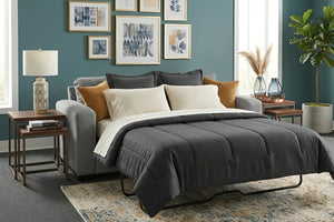 Sofá cama tapizado en tela gris convertido en cama matrimonial, con ropa de cama gris y almohadas decorativas. Ubicado en una sala moderna con mesas auxiliares, lámpara, cuadros en la pared y planta decorativa.