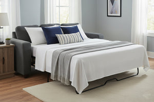 Sofá cama convertido en cama de tamaño completo con ropa de cama blanca y manta gris en una habitación con piso de madera y paredes gris claro.