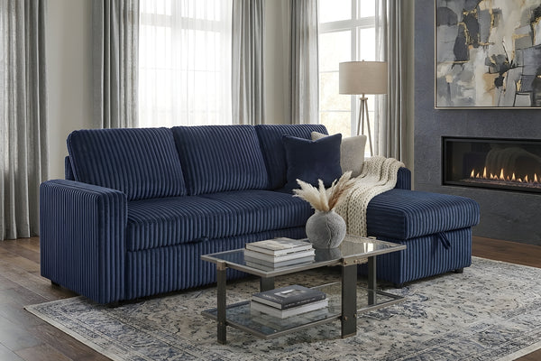 Sofá seccional reversible tapizado en tela acanalada azul marino, con chaise longue integrada y cojines decorativos. Se muestra en una sala moderna junto a una mesa de centro de vidrio, alfombra y chimenea.