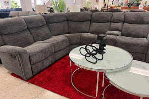 Sofá modular gris en un entorno de exposición con una mesa redonda de cristal y una alfombra roja. 