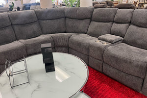Sofá modular gris en un entorno de exposición con una mesa redonda de cristal y una alfombra roja. 