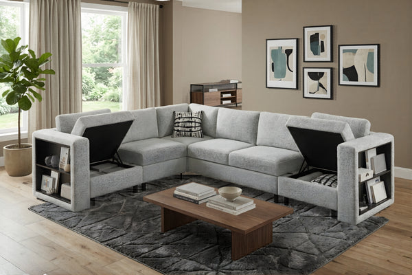 Sofá seccional modular en forma de U tapizado en tela gris clara con compartimentos de almacenamiento abiertos en ambos brazos y cojines elevables. El conjunto incluye cojines decorativos estampados, mesa de centro de madera y alfombra oscura en una sala contemporánea con arte abstracto y luz natural.