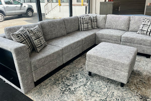 Sofá seccional en forma de L tapizado en tela gris jaspeada con cojines decorativos estampados en tonos negro y blanco, acompañado de un otomano cuadrado a juego. El conjunto está exhibido sobre una alfombra con diseño ornamental en una sala de exhibición moderna con ventanales amplios.