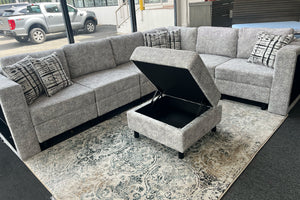 Sofá seccional en forma de L tapizado en tela gris jaspeada con cojines decorativos estampados y otomano cuadrado con compartimento de almacenamiento abierto. El conjunto está exhibido sobre una alfombra con diseño ornamental en una sala de exhibición con ventanales grandes.