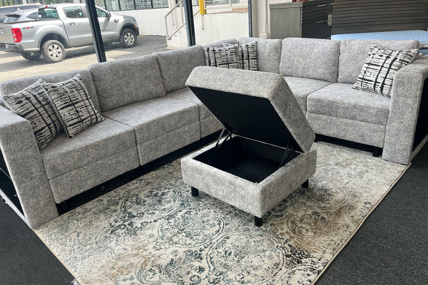 Sofá seccional en forma de L tapizado en tela gris jaspeada con cojines decorativos estampados y otomano cuadrado con compartimento de almacenamiento abierto. El conjunto está exhibido sobre una alfombra con diseño ornamental en una sala de exhibición con ventanales grandes.
