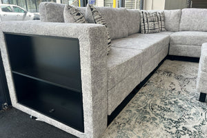 Vista lateral de un sofá seccional tapizado en tela gris jaspeada con compartimento de almacenamiento abierto integrado en el brazo lateral. El sofá incluye cojines decorativos estampados y está colocado sobre una alfombra con diseño ornamental en una sala de exhibición moderna.
