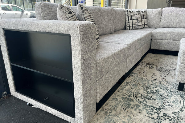 Vista lateral de un sofá seccional tapizado en tela gris jaspeada con compartimento de almacenamiento abierto integrado en el brazo lateral. El sofá incluye cojines decorativos estampados y está colocado sobre una alfombra con diseño ornamental en una sala de exhibición moderna.