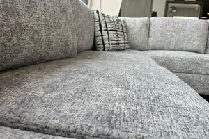 Primer plano de los cojines de asiento y respaldo de un sofá seccional tapizado en tela gris jaspeada, mostrando la textura suave del tapizado y cojines decorativos estampados en blanco y negro.