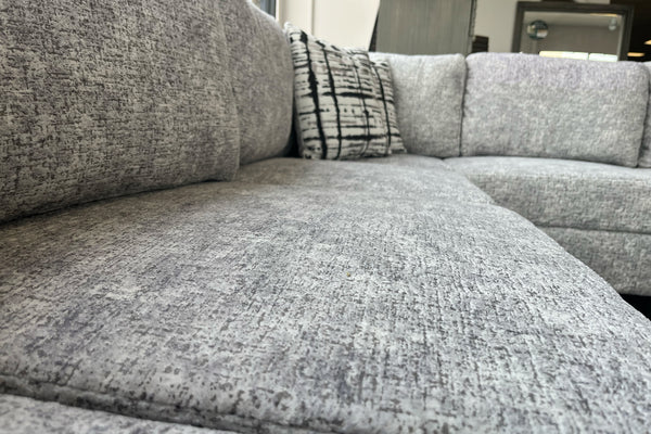 Primer plano de los cojines de asiento y respaldo de un sofá seccional tapizado en tela gris jaspeada, mostrando la textura suave del tapizado y cojines decorativos estampados en blanco y negro.