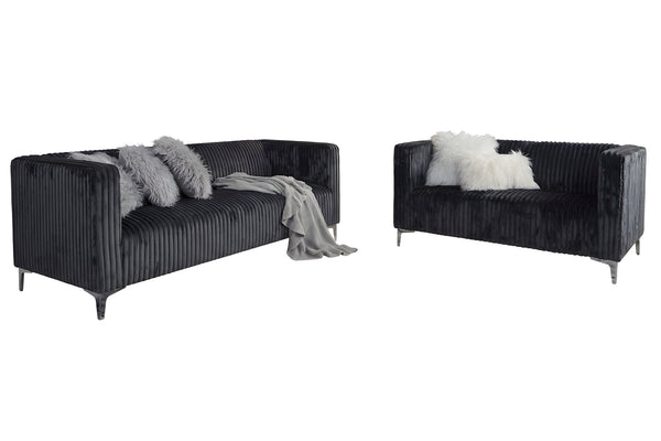 Sofá y loveseat de terciopelo gris oscuro con cojines decorativos sobre un fondo blanco.