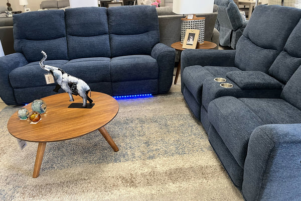 Juego de sala reclinable en tela azul oscuro con sofá y loveseat, respaldos acolchados, consola con portavasos y luces LED inferiores, exhibido junto a mesa de centro redonda de madera en una sala moderna.