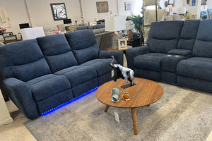 Sala reclinable en tela azul oscuro con sofá y loveseat acolchados, consola central con portavasos y luces LED azules en la base, acompañados de mesa de centro redonda de madera en showroom de muebles.