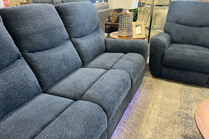 Primer plano de sofá reclinable en tela azul oscuro con cojines acolchados y diseño contemporáneo, acompañado de sillón a juego y detalles decorativos en showroom de muebles modernos.