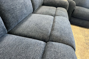 Primer plano de sofá reclinable en tela azul oscuro con textura tejida y cojines acolchados, mostrando asientos amplios y diseño cómodo en exhibición de tienda de muebles.