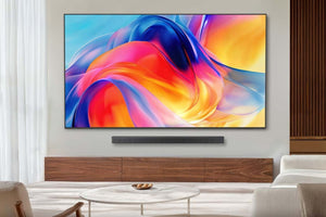Televisor de pantalla plana montado en pared con diseño delgado y marco negro minimalista, mostrando una imagen abstracta de colores vibrantes en tonos azul, rojo, naranja y amarillo. Debajo del televisor se encuentra una barra de sonido negra colocada sobre un mueble de madera de tono cálido. La escena está ambientada en una sala moderna con cortinas claras, una mesa de centro redonda y sofás en tonos neutros.