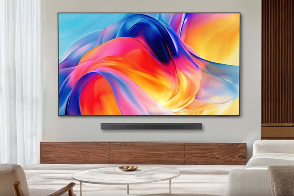 Televisor de pantalla plana montado en pared con diseño delgado y marco negro minimalista, mostrando una imagen abstracta de colores vibrantes en tonos azul, rojo, naranja y amarillo. Debajo del televisor se encuentra una barra de sonido negra colocada sobre un mueble de madera de tono cálido. La escena está ambientada en una sala moderna con cortinas claras, una mesa de centro redonda y sofás en tonos neutros.