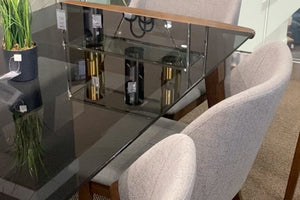 Mesa de comedor rectangular con cubierta de vidrio ahumado y estante central de cristal. Rodeada por sillas tapizadas en tono gris claro con brazos curvos. Diseño contemporáneo exhibido en una sala de muebles.