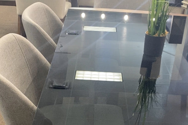 Mesa de comedor rectangular con cubierta de vidrio ahumado y acabado brillante, acompañada de sillas tapizadas en tono gris claro. La superficie refleja la iluminación superior y cuenta con un centro decorativo en maceta, resaltando su estilo moderno.