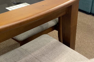 Detalle de mesa de comedor con estructura de madera en tono nogal y silla tapizada en tela gris claro. La imagen destaca los acabados de la madera, el diseño robusto de la base y la textura suave del asiento acolchado.