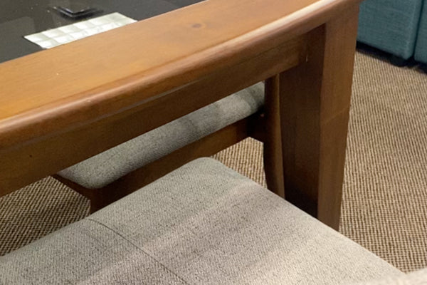 Detalle de mesa de comedor con estructura de madera en tono nogal y silla tapizada en tela gris claro. La imagen destaca los acabados de la madera, el diseño robusto de la base y la textura suave del asiento acolchado.