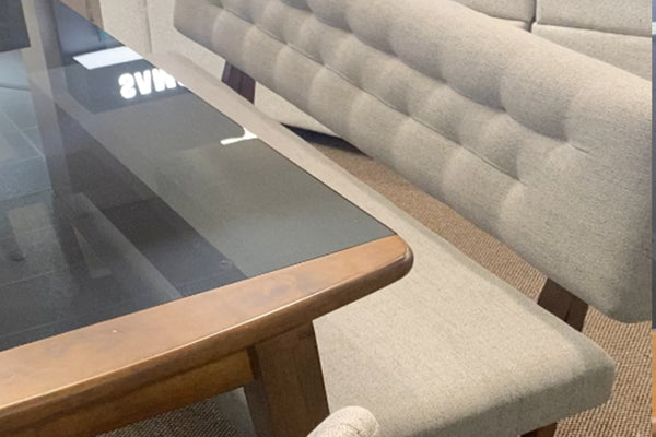 Detalle de juego de comedor con mesa rectangular de vidrio ahumado y marco de madera en tono nogal. Se aprecia un banco tapizado en tela gris con respaldo acolchado capitoné y asiento a juego, destacando sus texturas y acabados contemporáneos.