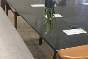 Mesa de comedor rectangular con cubierta de vidrio ahumado y estructura de madera en tono nogal. A un lado se observa un banco tapizado en tela gris clara. La imagen destaca la superficie brillante, los reflejos del vidrio y el diseño contemporáneo del conjunto.