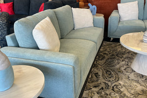 Sofá moderno tapizado en tela verde claro con cojines decorativos blancos, acompañado de sillón a juego y mesas redondas con acabado tipo mármol, ubicado sobre una alfombra estampada en una sala de exhibición contemporánea.