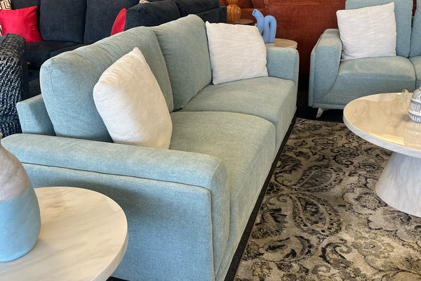Sofá moderno tapizado en tela verde claro con cojines decorativos blancos, acompañado de sillón a juego y mesas redondas con acabado tipo mármol, ubicado sobre una alfombra estampada en una sala de exhibición contemporánea.