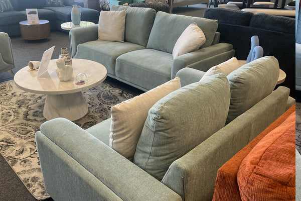 Juego de sala contemporáneo con sofá y loveseat tapizados en tela verde claro, cojines decorativos beige y mesa de centro redonda con acabado tipo mármol, presentado sobre una alfombra estampada en tonos grises dentro de una tienda de muebles.