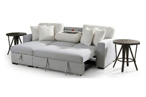 Sofá cama gris claro con cojines blancos acanalados, asientos extensibles tipo chaise y acompañado de dos mesas laterales oscuras.
