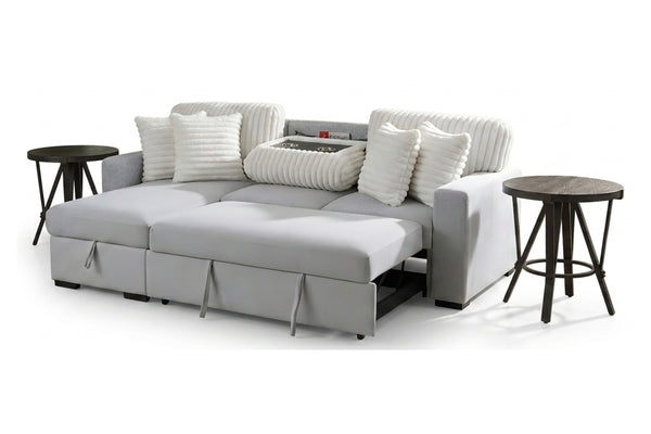 Sofá cama gris claro con cojines blancos acanalados, asientos extensibles tipo chaise y acompañado de dos mesas laterales oscuras.
