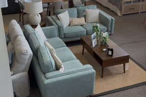 Vista superior de sala moderna con sofá y loveseat en tela verde agua, cojines decorativos claros y mesa de centro de madera oscura sobre alfombra beige en showroom de muebles contemporáneos.