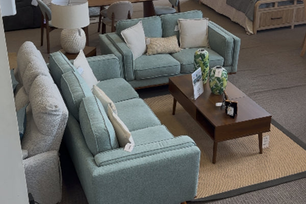 Vista superior de sala moderna con sofá y loveseat en tela verde agua, cojines decorativos claros y mesa de centro de madera oscura sobre alfombra beige en showroom de muebles contemporáneos.