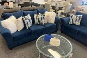 Juego de sala en terciopelo azul marino con sofá y sillón acolchados, decorados con cojines estampados y peludos blancos, acompañado de mesa de centro redonda de vidrio en showroom de muebles modernos.