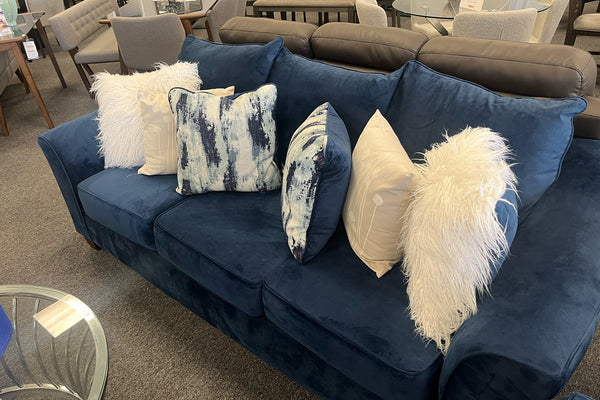 Sofá en terciopelo azul marino con cojines decorativos blancos y estampados en tonos azules, respaldo acolchado y diseño contemporáneo, exhibido en showroom de muebles modernos.