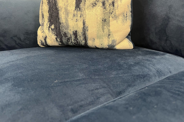 Primer plano de asiento en terciopelo azul marino con cojín decorativo estampado en tonos crema y azul, resaltando la textura suave y acolchada del sofá moderno.