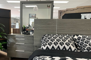 Vista cercana de juego de dormitorio moderno en acabado gris en tienda, con cabecera de paneles, ropa de cama en blanco y negro con patrón geométrico, y gavetero con tiradores metálicos y espejo rectangular encima.