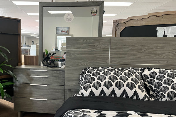 Vista cercana de juego de dormitorio moderno en acabado gris en tienda, con cabecera de paneles, ropa de cama en blanco y negro con patrón geométrico, y gavetero con tiradores metálicos y espejo rectangular encima.
