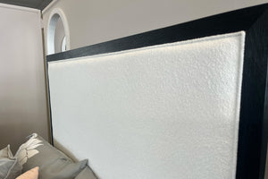 Detalle de cabecera de cama tapizada en tela beige con textura suave y marco de madera oscura en la parte superior y laterales. Incluye almohadas en tonos neutros y se muestra en un entorno interior con pared clara y arco en el fondo.