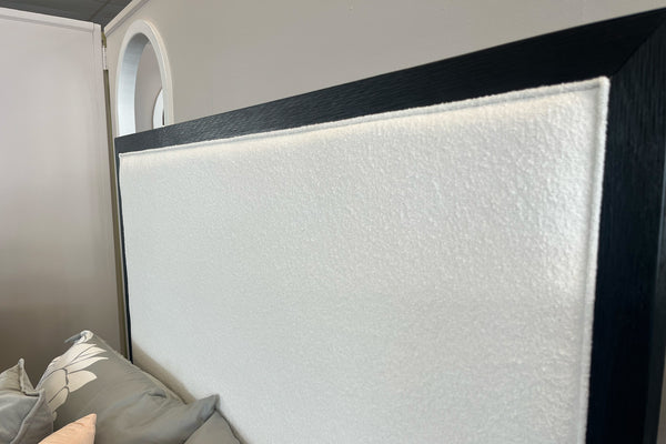 Detalle de cabecera de cama tapizada en tela beige con textura suave y marco de madera oscura en la parte superior y laterales. Incluye almohadas en tonos neutros y se muestra en un entorno interior con pared clara y arco en el fondo.