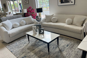 Sala moderna con sofá y loveseat curvos en tela color marfil, cojines decorativos redondos y mesa de centro blanca sobre alfombra estampada, exhibidos en showroom de muebles contemporáneos.