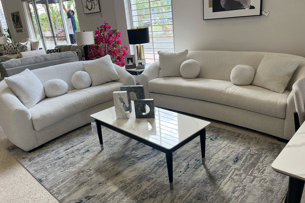 Sala moderna con sofá y loveseat curvos en tela color marfil, cojines decorativos redondos y mesa de centro blanca sobre alfombra estampada, exhibidos en showroom de muebles contemporáneos.
