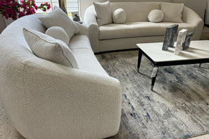 Sofás curvos en tela color marfil con cojines decorativos redondos y cuadrados, acompañados de mesa de centro blanca sobre alfombra estampada en una sala moderna y elegante.