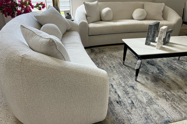 Sofás curvos en tela color marfil con cojines decorativos redondos y cuadrados, acompañados de mesa de centro blanca sobre alfombra estampada en una sala moderna y elegante.